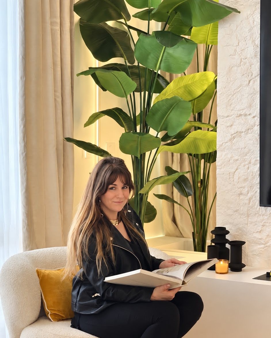 Déborah, fondatrice de PsychéDéco, assise dans un fauteuil crème, un livre de décoration ouvert sur les genoux